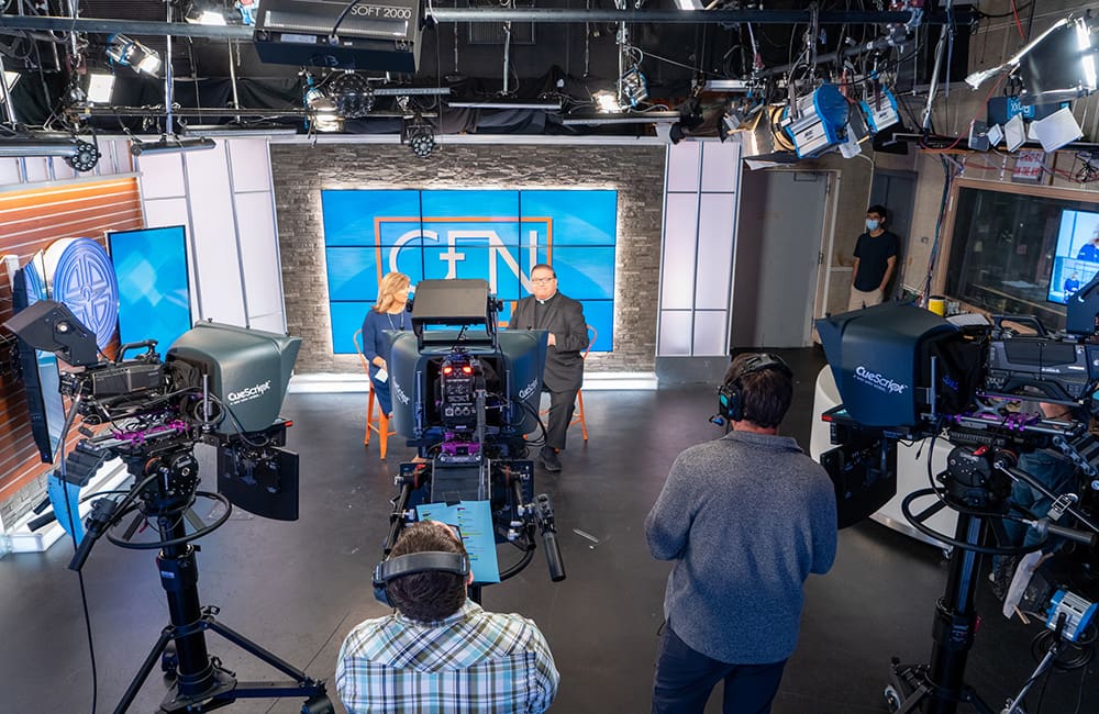 A television studio with two men and one man is standing in front of the camera.